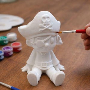 May include: A white ceramic pirate figurine with a hat, eye patch, and skull and crossbones detail. A person is painting the figurine with a red paintbrush. Paint pots and a blue paintbrush are also on the wooden surface.