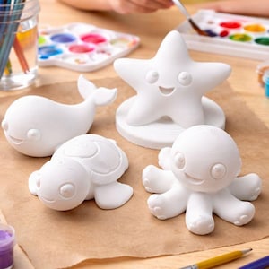 May include: White ceramic sea creature figurines, including a starfish, whale, turtle, and octopus, are arranged on a craft table. Paintbrushes, paint palettes, and small paint pots in various colors are also present, suggesting a DIY painting activity.
