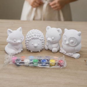 May include: A set of four white ceramic animal figurines including a fox, a hedgehog, a bear, and an owl. The set includes a plastic strip of twelve small paint pots in various colors and several paintbrushes in a glass jar and on the table.
