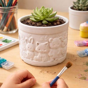 May include: A white ceramic planter with embossed animal designs, including a rabbit, cat, and bear, holds a green succulent. A paintbrush with a blue handle is held by a person. Other art supplies are visible.