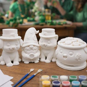 May include: A collection of unpainted ceramic figurines, including a dog, a gnome, a cat, and a pot of gold, arranged on a wooden table. Paintbrushes and a set of colorful paints are also present, suggesting a DIY painting activity. The background shows a St. Patrick's Day celebration.
