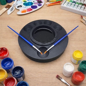 Pode incluir: Uma coleção de materiais de arte, incluindo pincéis, tubos de tinta e potes de tinta em várias cores. Uma paleta de artista preta com dois pincéis azuis está no centro. Uma paleta de plástico branca com cores de tinta misturadas também é visível.