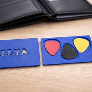 May include: Black leather wallet open on a wooden surface, with a blue holder containing three guitar picks in red, black, and yellow. A blue card with the cut-out text "ILYA" is also present.