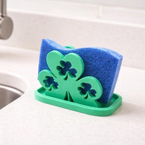 May include: A green shamrock-shaped sponge holder with a blue sponge. The holder is made of plastic and sits on a countertop next to a stainless steel sink and faucet. The shamrock design is a prominent feature.