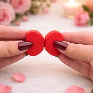 May include: A bright red bow-shaped object is held between two hands with dark red nail polish. The background features soft pink roses, candles, and bokeh lights, creating a romantic ambiance. The bow appears to be made of a soft material.