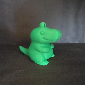 Peut inclure: Une figurine de dinosaure vert vif avec une boucle sur le dessus. Le dinosaure de dessin animé a un visage souriant, de petits bras et une queue. La figurine est faite d'un matériau solide et repose sur une surface sombre.