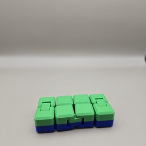 Puede incluir: Un juguete fidget verde y azul con un dise&ntilde;o segmentado. El juguete est&aacute; compuesto por varios bloques conectados, cada uno con una parte superior verde y una base azul. Los bloques est&aacute;n dispuestos de forma lineal, lo que permite una manipulaci&oacute;n flexible.