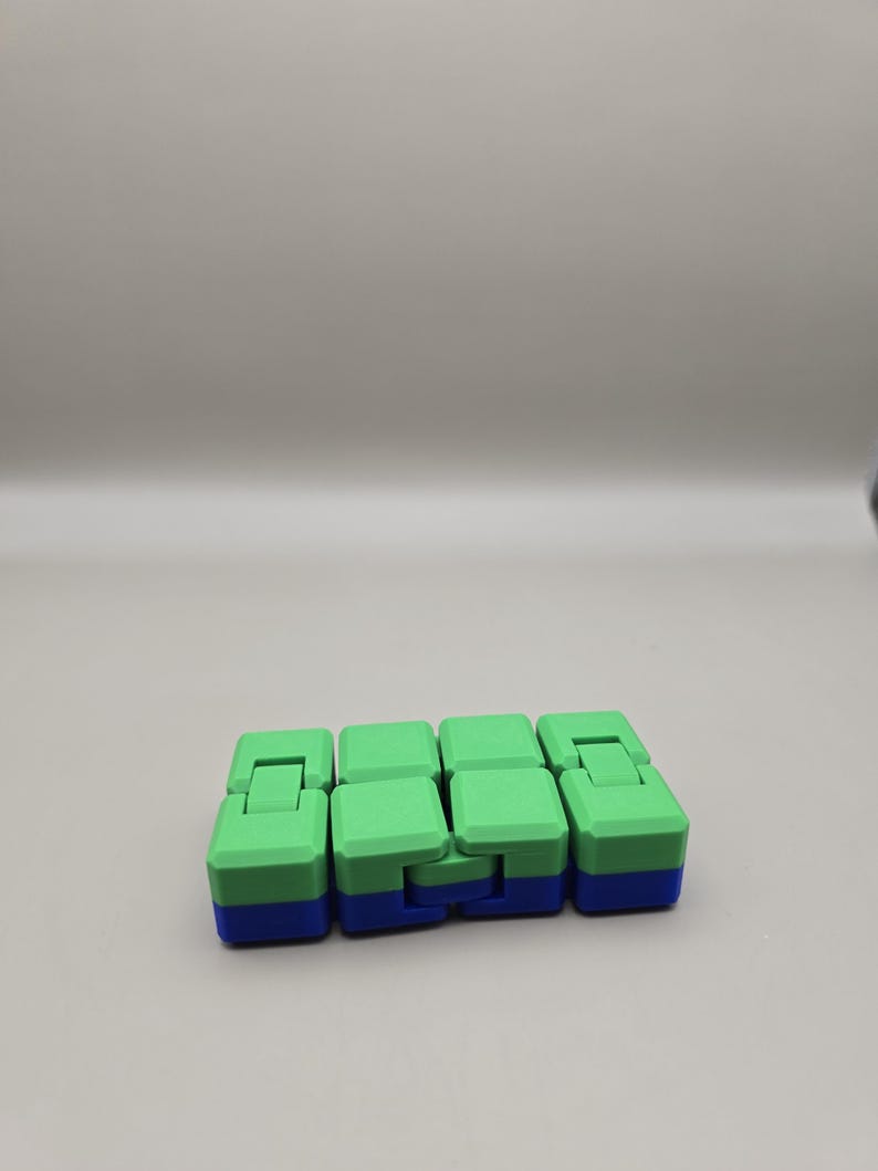 Puede incluir: Un juguete fidget verde y azul con un dise&ntilde;o segmentado. El juguete est&aacute; compuesto por varios bloques conectados, cada uno con una parte superior verde y una base azul. Los bloques est&aacute;n dispuestos de forma lineal, lo que permite una manipulaci&oacute;n flexible.