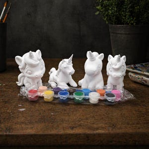 May include: Four white unicorn figurines in various poses, ready to be painted, sit on a wooden surface. A small paint set with multiple colors is in front of the unicorns. Paintbrushes in a black container are in the background.