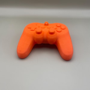 Op de afbeelding: Fel oranje videogamecontroller met knoppen en handvatten. De controller heeft een lus aan de bovenkant. De controller heeft een effen kleur en ligt op een grijze ondergrond.