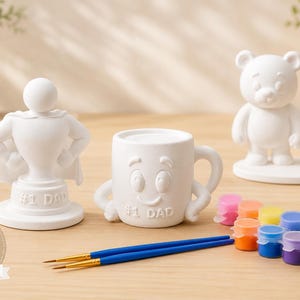 May include: A collection of white ceramic Father's Day gifts, including a superhero trophy, a smiling mug, and a bear family figurine. Paintbrushes and paint pots in various colors are also included. The words "Paint Create Celebrate" and "Father's Day" are visible.