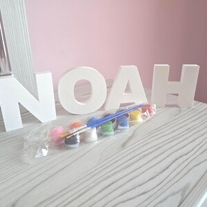 May include: White wooden letters spelling "NOAH" are arranged on a gray wooden surface. A paint set with brushes and colorful paints is in front of the letters. The background is a light pink wall.
