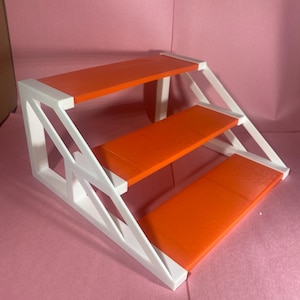 May include: A three-tiered display stand with an orange surface and white supports. The stand has a stepped design, with each level decreasing in size. The stand is made of a solid material and is suitable for displaying small items.