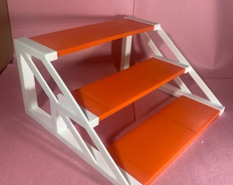 May include: A three-tiered display stand with an orange surface and white supports. The stand has a stepped design, with each level decreasing in size. The stand is made of a solid material and is suitable for displaying small items.