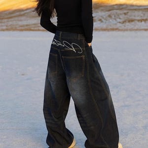 May include: Dark wash denim jeans with a wide leg design. The jeans have a high waist and feature white embroidered detailing on the back pockets. The jeans are paired with a black long-sleeve top and white trainers.