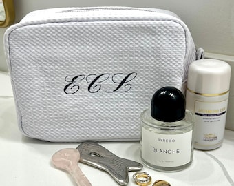 May include: White toiletry bag with the initials "ECL" embroidered on the front. The bag is next to a bottle of Byredo Blanche perfume, a rose quartz gua sha tool, and a silver facial tool. There are also two gold rings.
