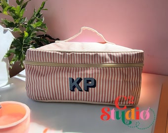 May include: Pink and white striped fabric cosmetic bag with a zipper closure and a navy blue embroidered monogram "KP".