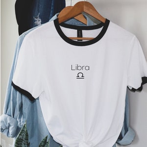 Puede incluir: Camiseta blanca con un ribete negro y el texto "Libra" y el s&iacute;mbolo del zod&iacute;aco de Libra en negro.