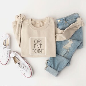 May include: A beige sweatshirt with the text "ORIENT POINT" in a black box on the front. The sweatshirt is laid out on a white surface with a pair of white canvas shoes and a pair of light blue jeans.