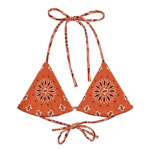 May include: A rust-colored bikini top with a bandana print. The triangle cups feature a central floral design in black and white, with matching tie straps at the neck and back. The overall design is a classic, bohemian style.