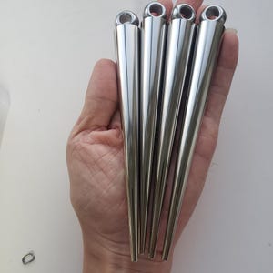 Puede incluir: Cuatro puntas de metal plateado pulido, cada una con una forma cónica lisa y afilada, y un bucle circular en el extremo superior más ancho. Estos objetos metálicos están dispuestos uno al lado del otro con un acabado brillante.