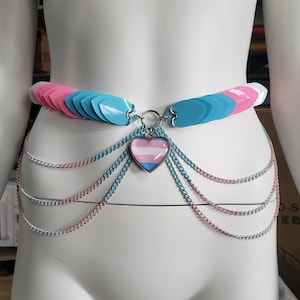 Puede incluir: Un cinturón de cadena azul, rosa y blanco con un dije de corazón de orgullo transgénero. El cinturón está hecho de escamas de plástico y cadenas metálicas.