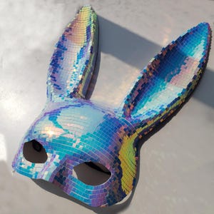 May include: A decorative bunny mask with iridescent, mosaic-like tiles. The mask features long, upright ears and eye openings. The color shifts between blue, purple, and green, creating a shimmering effect. The mask is designed for costume or party use.