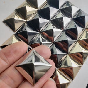 Könnte beinhalten: Eine Sammlung von metallischen pyramidenförmigen Nieten mit reflektierender Oberfläche. Die Nieten sind in einem Gittermuster angeordnet und weisen abwechselnd goldene und silberne Töne auf. Jede einzelne Niete hat vier dreieckige Flächen.