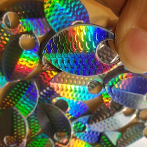 Peut inclure: Gros plan sur plusieurs leurres de pêche irisés. Les leurres en forme de goutte d'eau présentent une texture en écailles, reflétant un arc-en-ciel de couleurs. Chaque leurre est percé d'un petit trou pour l'attache.