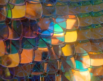May include: Close-up of iridescent blue, green, and gold sequins arranged in a scaly pattern. The sequins are overlapping and create a shimmering effect.