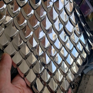 Puede incluir: Una pieza de armadura de escamas con escamas metálicas superpuestas de color plateado pulido, dispuestas en un patrón uniforme y repetitivo. Cada escama tiene una forma puntiaguda similar a un escudo, creando una superficie reflectante y texturizada.