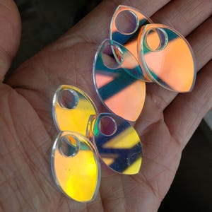 Puede incluir: Un conjunto de cinco escamas iridiscentes en forma de lágrima con una superficie lisa y reflectante. Cada escama presenta un orificio circular en la parte superior, mostrando un espectro de colores cambiantes que incluyen tonos naranja, amarillo y azul.