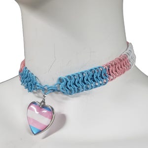 May include: A chainmail choker necklace with a heart pendant. The choker features blue, pink, and white chainmail links. The heart pendant has a silver border and displays the transgender pride flag.