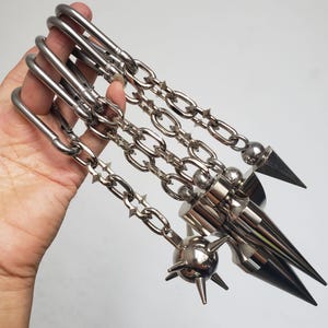 Puede incluir: Un conjunto de tres cadenas de metal plateado, cada una unida a un mosquetón. Las cadenas presentan pequeñas púas en los eslabones y terminan en pesados colgantes de metal, incluyendo una bola esférica con púas y dos conos lisos y puntiagudos.