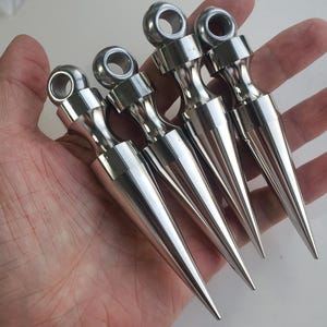 Puede incluir: Cuatro puntas de metal plateado pulido, cada una con un cuerpo cónico y un ojal circular en la parte superior. Estos componentes metálicos tienen un acabado liso y reflectante y están dispuestos en una fila paralela, mostrando sus puntas.