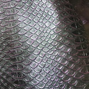 Puede incluir: Primer plano de un material plateado metálico texturizado con un patrón de escamas. Las escamas tienen un brillo sutil e iridiscente, con toques de morado y rosa. La superficie parece ser cuero o un material similar.