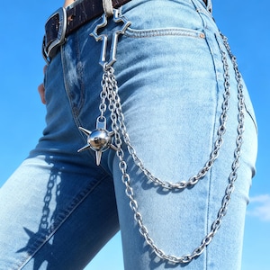 May include: Silver-toned chain belt with a cross-shaped top piece and a spiked ball pendant. The chain hangs down the front of light blue denim jeans. The belt is worn with a brown leather belt and a silver buckle.