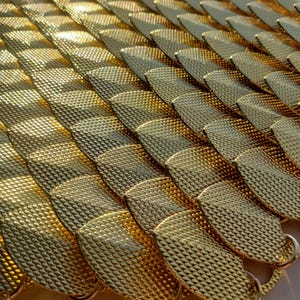 May include: Close-up of a golden, textured material arranged in overlapping, scale-like patterns. The surface reflects light, creating highlights and shadows. The design suggests a decorative or ornamental element, possibly for fashion or craft projects.