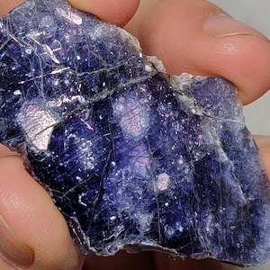 以下が含まれることがあります： 手で持たれた、生の虹色の青紫色の蛍石の結晶。石は、内部構造が見え、キラキラと反射する表面を持つ、割れた外観をしています。色は濃いインディゴから明るい色合いに変化します。
