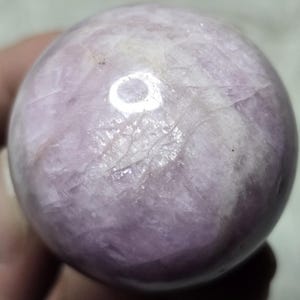May include: A polished, spherical gemstone in shades of lavender and white. The crystal ball has a smooth surface with natural veining patterns. The sphere is held in a person's hand.