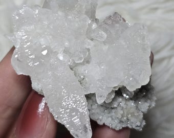 Op de afbeelding: Een cluster van heldere kwartskristallen. De kwartskristallen hebben een doorschijnende witte kleur en zijn bevestigd aan een grijze, getextureerde basis. De kristallen hebben scherpe, puntige uiteinden.