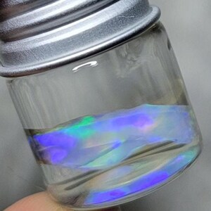 May include: A small, clear glass jar with a silver-colored metal screw-top lid. Inside the jar, there is a layer of iridescent material that reflects vibrant shades of blue, purple, and green, creating a shimmering, holographic effect within the container.