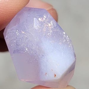 Puede incluir: Un cristal facetado, de color púrpura claro, sostenido entre dos dedos. El cristal tiene una cualidad translúcida con tonos blancos y lavanda. La forma del cristal es irregular, con múltiples facetas que captan la luz. La apariencia general es suave y pulida.