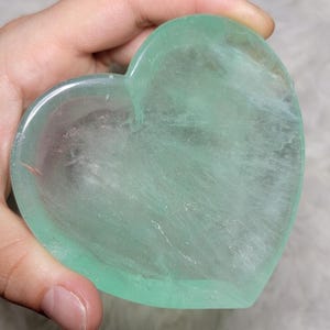 Può includere: Un cristallo di fluorite verde lucidato a forma di cuore con qualità traslucide e inclusioni minerali interne visibili. La pietra presenta una superficie liscia e arrotondata con varie tonalità di verde menta e verde pallido.