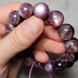 May include: A beaded bracelet featuring round, polished purple stones with a chatoyant, shimmering effect. The beads are strung on a thin, light purple elastic cord, displaying natural variations in color and light reflection across the surface.