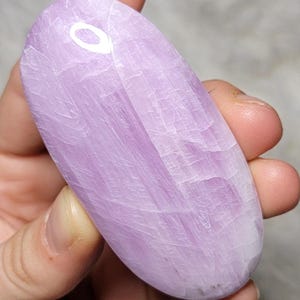 May include: An oval-shaped, polished lavender-colored stone with a smooth, glossy surface and visible internal crystalline fractures. The stone displays varying shades of light purple and white, with a distinct reflective highlight on its upper portion.