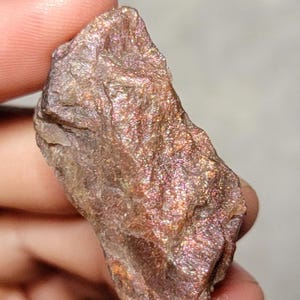 May include: A rough-cut, iridescent sunstone gemstone. The stone displays a mix of brown, orange, and pink hues with a shimmering, glittery effect. The surface has a textured, uneven appearance, showcasing natural mineral formations.