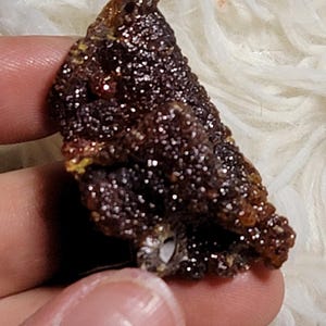 Puede incluir: Un espécimen mineral de color marrón oscuro con una estructura cristalina, sostenido en una mano. La superficie brilla con pequeños cristales reflectantes. El mineral tiene una apariencia rugosa y texturizada y una forma ligeramente redondeada.