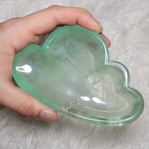 Può includere: Una ciotola in cristallo di fluorite verde traslucido e levigato, intagliata a forma di nuvola. Il minerale presenta inclusioni naturali, sfumature dal verde chiaro al medio e una superficie liscia e lucida con riflessi luminosi.