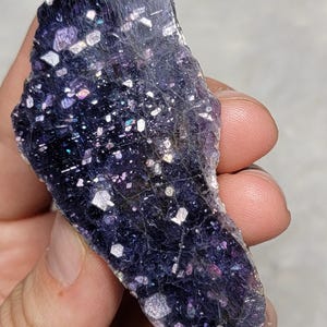 以下が含まれることがあります： 暗い紫色の、不規則な形の石で、虹色の結晶状のインクルージョンがあります。石は手で持たれており、その自然な質感と色のバリエーションを示しています。表面は白と銀のフレークで輝いています。